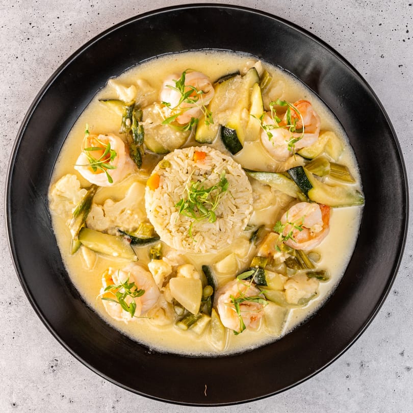 Overhead view of a creamy shrimp and vegetable dish with a mound of rice in a black bowl.