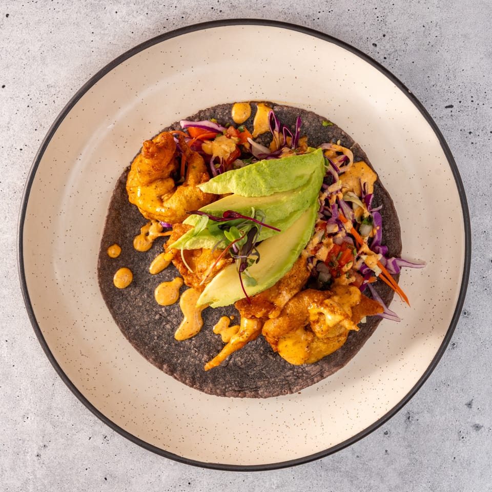 A Raices Cocina & Bar shrimp taco with avocado, cabbage, and sauce on a blue corn tortilla.