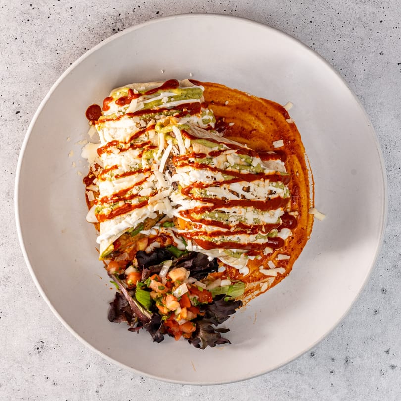 Mexican enchiladas with red sauce, green sauce, white cheese, and pico de gallo on a white plate.
